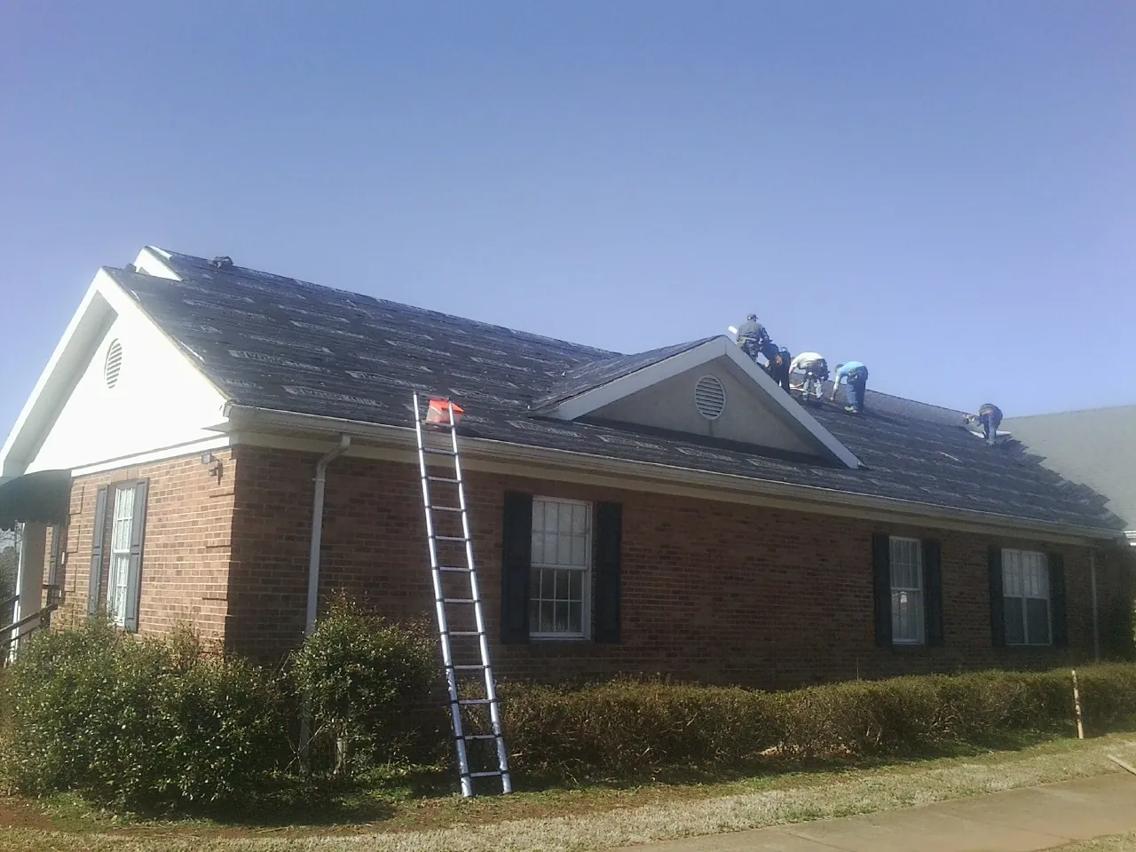 Crew tearing shingles off a brick ranch roof with a ladder set — Metal Roof Repair work by our crew in Elma