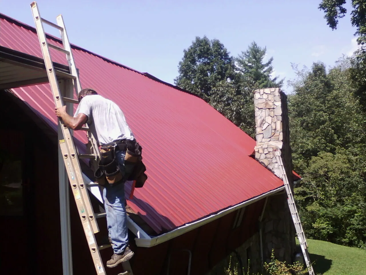 Roofer on a ladder beside a red metal roof and stone chimney — Soffit & Fascia Repair by Roofing Brothers in Elma